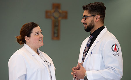 A female and male pharmacy student in lab coats talking in front a wall with a cross.